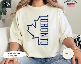 May include: A white t-shirt with a blue maple leaf and the word "TORONTO" printed vertically on the front. The shirt is being modeled by a person wearing blue jeans. The text "IVORY" is visible in the lower right corner of the image.