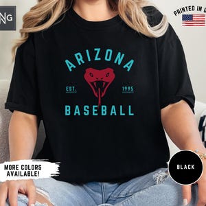 以下が含まれることがあります： 黒いTシャツ。ターコイズ色の「ARIZONA BASEBALL」の文字と赤いヘビのグラフィック。シャツには「EST. 1995」の文字も含まれています。「PRINTED IN USA」の文字とアメリカ国旗が見えます。