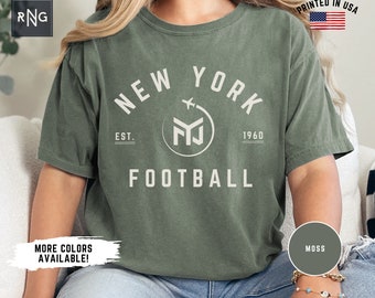 Könnte beinhalten: Ein moosgrünes T-Shirt mit einem weißen Grafikdruck, auf dem "New York" in einem Bogen mit einem Flugzeug steht, das über den Buchstaben "Y" und "O" fliegt. Die Grafik enthält auch einen Kreis mit den Buchstaben "NY" darin und den Text "Est. 1960" und "Football" unter dem Bogen.