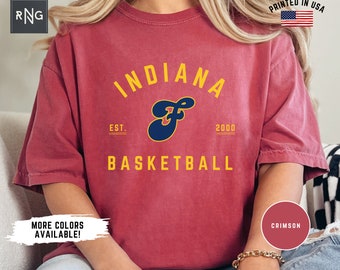 May include: Crimson red t-shirt with a vintage-style Indiana University basketball logo. The logo features a blue "F" with a yellow outline and the text "Indiana" in yellow above and "Basketball" in yellow below. The text "Est. 2000" is in yellow below the logo.