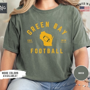 Peut inclure: Un t-shirt &agrave; manches courtes vert mousse avec un texte jaune indiquant GREEN BAY FOOTBALL et EST. 1919. Le motif comprend une silhouette jaune de l'&Eacute;tat du Wisconsin avec un ballon de football au centre. Le t-shirt a une encolure ronde.