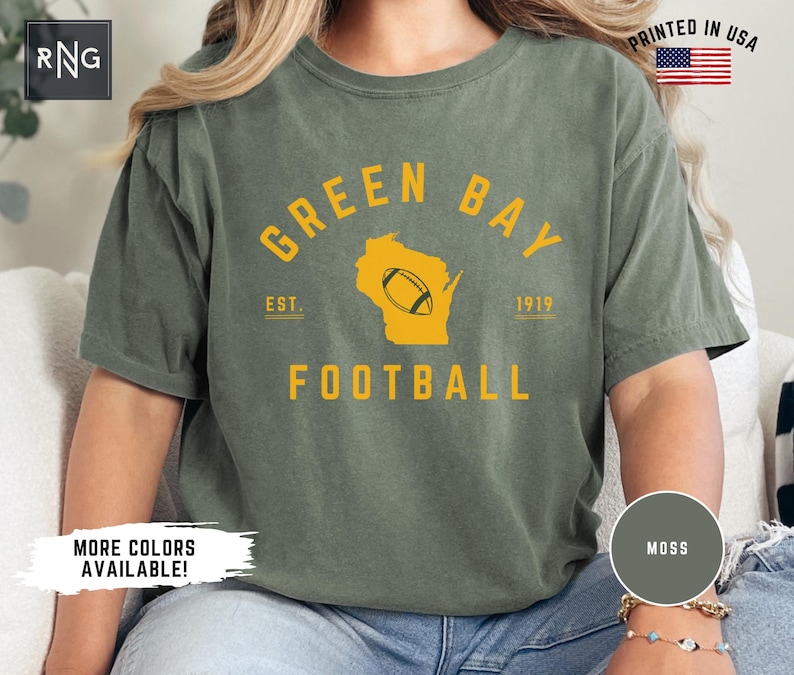 Peut inclure: Un t-shirt &agrave; manches courtes vert mousse avec un texte jaune indiquant GREEN BAY FOOTBALL et EST. 1919. Le motif comprend une silhouette jaune de l'&Eacute;tat du Wisconsin avec un ballon de football au centre. Le t-shirt a une encolure ronde.
