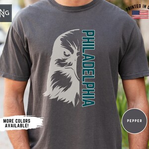 K&ouml;nnte beinhalten: Ein dunkelgraues T-Shirt mit einem wei&szlig;en und t&uuml;rkisfarbenen Adler-Motiv und dem Wort "PHILADELPHIA", das vertikal auf der rechten Seite des Shirts aufgedruckt ist.