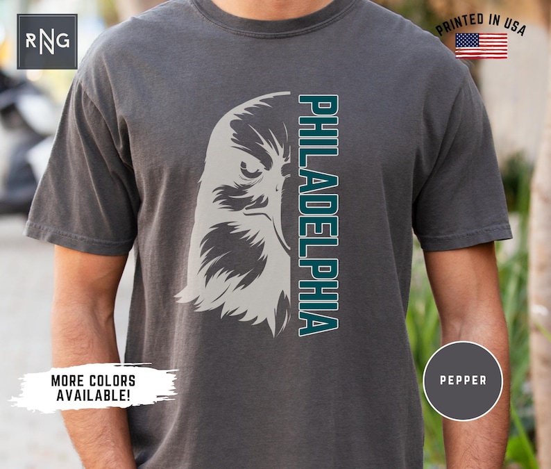 K&ouml;nnte beinhalten: Ein dunkelgraues T-Shirt mit einem wei&szlig;en und t&uuml;rkisfarbenen Adler-Motiv und dem Wort "PHILADELPHIA", das vertikal auf der rechten Seite des Shirts aufgedruckt ist.