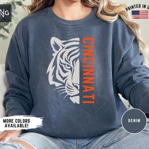 Puede incluir: Una sudadera azul marino con una cara de tigre blanca y la palabra "CINCINNATI" en letras naranjas. La sudadera est&aacute; hecha de un material suave y c&oacute;modo y tiene un corte holgado.