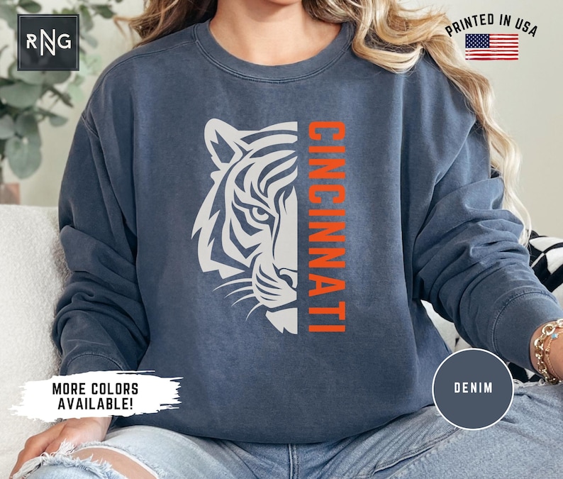 Puede incluir: Una sudadera azul marino con una cara de tigre blanca y la palabra "CINCINNATI" en letras naranjas. La sudadera est&aacute; hecha de un material suave y c&oacute;modo y tiene un corte holgado.