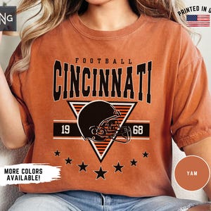 May include: A rust-colored short-sleeve t-shirt featuring a graphic print with the text FOOTBALL CINCINNATI, a football helmet, the numbers 19 and 68, and a row of stars. The design is centered on the chest in black and orange ink.