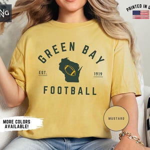 Peut inclure: Un t-shirt &agrave; manches courtes jaune moutarde avec un texte vert fonc&eacute; indiquant GREEN BAY FOOTBALL, EST. 1919, et une silhouette vert fonc&eacute; de l'&Eacute;tat du Wisconsin avec un graphique de ballon de football jaune au centre du motif.