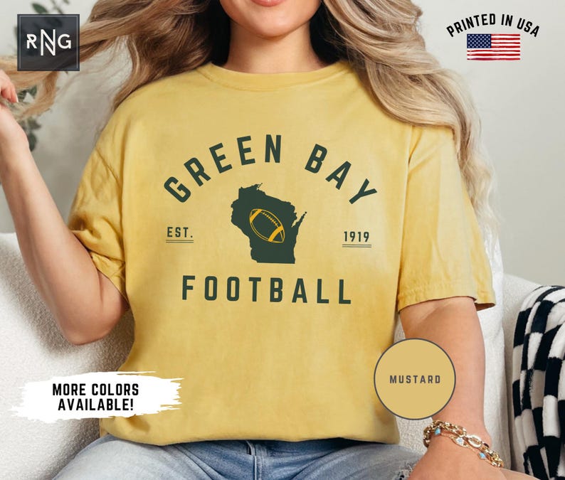 Peut inclure: Un t-shirt &agrave; manches courtes jaune moutarde avec un texte vert fonc&eacute; indiquant GREEN BAY FOOTBALL, EST. 1919, et une silhouette vert fonc&eacute; de l'&Eacute;tat du Wisconsin avec un graphique de ballon de football jaune au centre du motif.