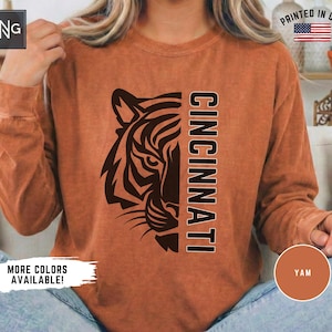 Könnte beinhalten: Ein langärmeliges Sweatshirt mit Rundhalsausschnitt in verwaschenem Rostorange. Auf der Vorderseite befindet sich links eine schwarze Grafik eines Tigerkopfes und rechts das vertikal in schwarzen Blockbuchstaben gedruckte Wort CINCINNATI.
