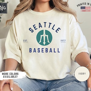 May include: A white t-shirt with a navy blue graphic of a trident logo and the text "SEATTLE EST. 1977 BASEBALL". The shirt is on a model wearing blue jeans. The text "MORE COLORS AVAILABLE!" is in the lower left corner and "IVORY" is in the lower right corner.