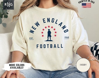 May include: Ivory t-shirt with a navy blue and red graphic of a person standing in front of a circle of stars with the text "NEW ENGLAND EST. 1960 FOOTBALL".