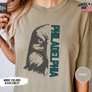 K&ouml;nnte beinhalten: Ein khakifarbenes T-Shirt mit einer Grafik eines stilisierten Adlerkopfes und dem Wort "PHILADELPHIA", das vertikal an der Seite aufgedruckt ist. Das Auge des Adlers ist dunkelgrau, und das Wort "PHILADELPHIA" ist in einem blaugr&uuml;nen Farbton gehalten. Das T-Shirt hat einen Used-Look und ist in den USA gedruckt.