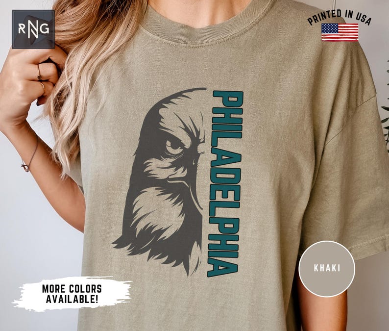 K&ouml;nnte beinhalten: Ein khakifarbenes T-Shirt mit einer Grafik eines stilisierten Adlerkopfes und dem Wort "PHILADELPHIA", das vertikal an der Seite aufgedruckt ist. Das Auge des Adlers ist dunkelgrau, und das Wort "PHILADELPHIA" ist in einem blaugr&uuml;nen Farbton gehalten. Das T-Shirt hat einen Used-Look und ist in den USA gedruckt.