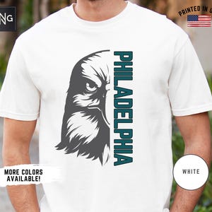K&ouml;nnte beinhalten: Wei&szlig;es T-Shirt mit einem Grafikdruck eines grauen Adlerkopfes und dem Wort "PHILADELPHIA" in t&uuml;rkisfarbenem Text.