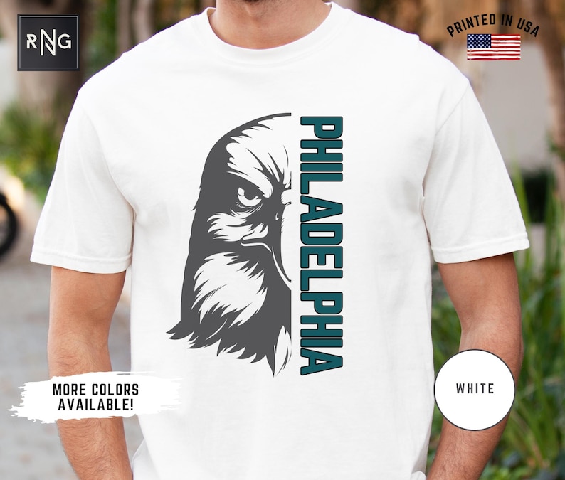 K&ouml;nnte beinhalten: Wei&szlig;es T-Shirt mit einem Grafikdruck eines grauen Adlerkopfes und dem Wort "PHILADELPHIA" in t&uuml;rkisfarbenem Text.
