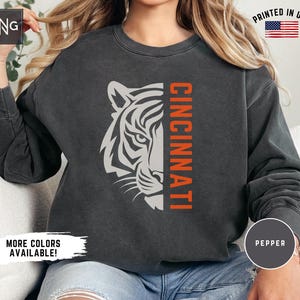 Puede incluir: Una sudadera negra con un gr&aacute;fico de tigre blanco y la palabra "CINCINNATI" en texto naranja. La sudadera est&aacute; hecha de un material suave y c&oacute;modo y tiene un aspecto vintage.