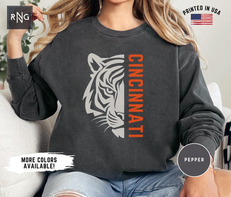 Puede incluir: Una sudadera negra con un gr&aacute;fico de tigre blanco y la palabra "CINCINNATI" en texto naranja. La sudadera est&aacute; hecha de un material suave y c&oacute;modo y tiene un aspecto vintage.
