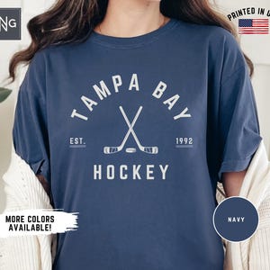 May include: A navy blue t-shirt with a white graphic of crossed hockey sticks and a puck. The graphic also includes the text "Tampa Bay", "Est. 1992", and "Hockey".