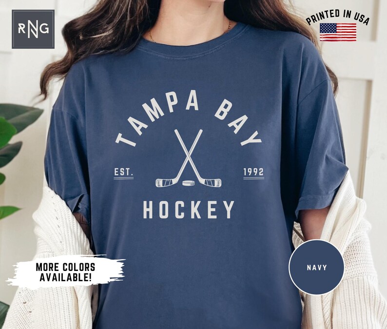 May include: A navy blue t-shirt with a white graphic of crossed hockey sticks and a puck. The graphic also includes the text "Tampa Bay", "Est. 1992", and "Hockey".