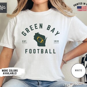 Peut inclure: Un t-shirt blanc &agrave; manches courtes avec un motif vert fonc&eacute; portant le texte GREEN BAY FOOTBALL, EST. 1919, et une silhouette de l'&Eacute;tat du Wisconsin contenant un ballon de football jaune. Le t-shirt pr&eacute;sente un logo RNG et une &eacute;tiquette PRINTED IN USA.