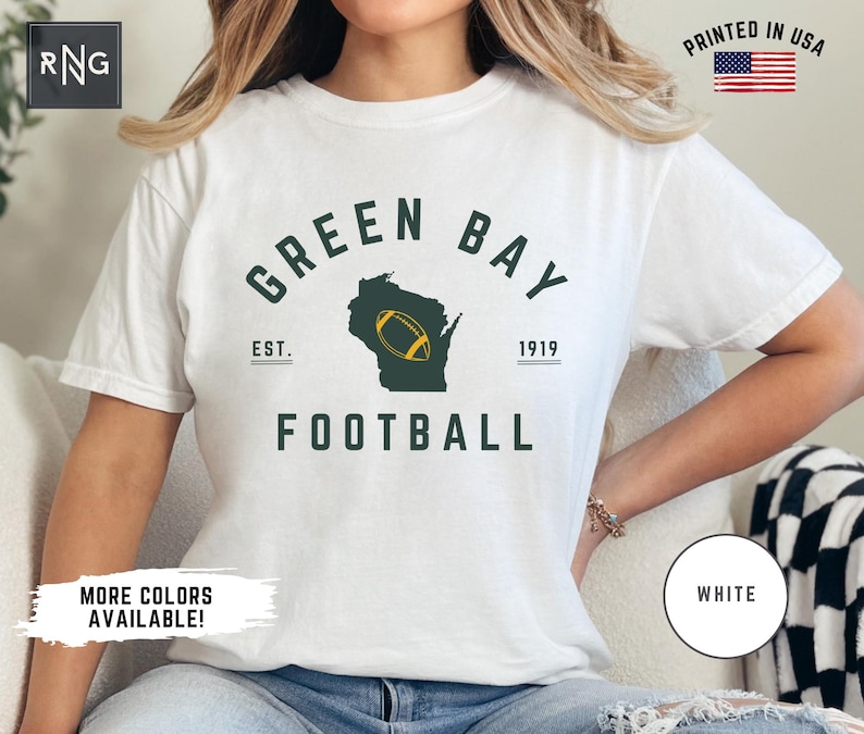 Peut inclure: Un t-shirt blanc &agrave; manches courtes avec un motif vert fonc&eacute; portant le texte GREEN BAY FOOTBALL, EST. 1919, et une silhouette de l'&Eacute;tat du Wisconsin contenant un ballon de football jaune. Le t-shirt pr&eacute;sente un logo RNG et une &eacute;tiquette PRINTED IN USA.