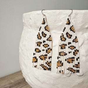 May include: A pair of beaded earrings with a leopard print design. The earrings are white with brown and black spots, and have a fringe at the bottom. The earrings are hanging on a white, textured surface.