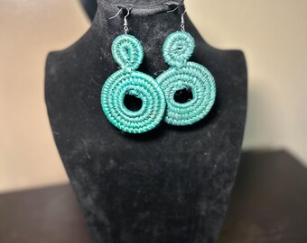 May include: A pair of turquoise woven earrings. Each earring features two connected circular shapes, with a smaller circle above a larger one. The earrings are displayed on a black mannequin head.