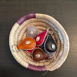 Puede incluir: Un conjunto de seis llaveros de cuero en una cesta tejida. Los llaveros tienen forma de lágrima y tienen un pequeño amuleto de concha blanca unido a cada uno. Los llaveros son de varios colores, incluyendo rojo, naranja, marrón, negro y gris.