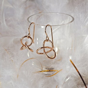 May include: A pair of gold-toned dangle earrings with a knotted design. The earrings are displayed in a glass container on a white feathery surface.