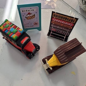 May include: A vintage toy set featuring two red and yellow toy cars and three miniature signs. The signs advertise ice cream sundaes, hamburgers, tenderloins, onion rings, root beer floats, and hot coffee.