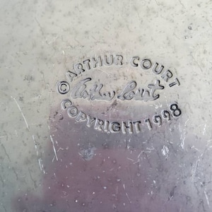 May include: Close-up of a silver metal surface with the text "Arthur Court Copyright 1998" and a handwritten signature.