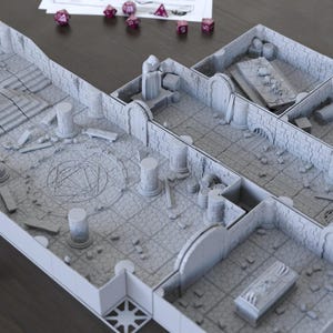 Op de afbeelding: Een grijs, modulair kerkerterrein voor tabletop gaming. De set bevat verschillende kamers, pilaren, trappen en een centraal cirkelvormig ontwerp. Er liggen verschillende dobbelstenen verspreid rond de set, wat op gameplay duidt. De set is ontworpen voor rollenspellen.