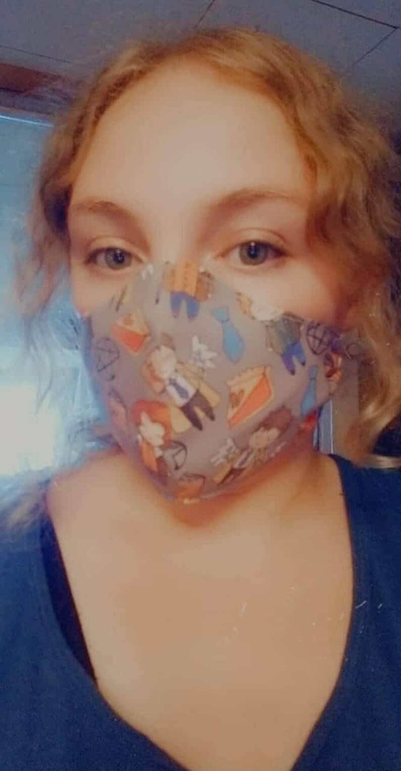 May include: A person wearing a gray face mask with a colorful pattern of cartoon characters and pie slices. The mask has a light gray background with a variety of colors, including blue, red, yellow, and green.