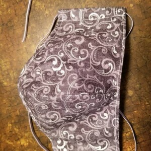 May include: A purple face mask with a white paisley pattern. The mask has elastic ear loops.