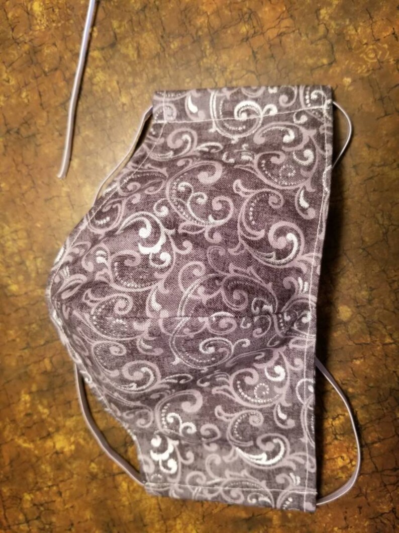 May include: A purple face mask with a white paisley pattern. The mask has elastic ear loops.