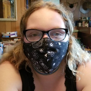 May include: A person wearing a black bandana face mask with a white paisley pattern. The person is wearing glasses and has long blonde hair.