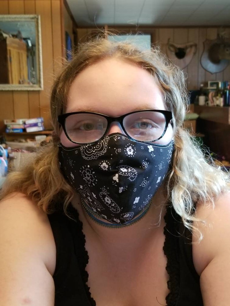 May include: A person wearing a black bandana face mask with a white paisley pattern. The person is wearing glasses and has long blonde hair.