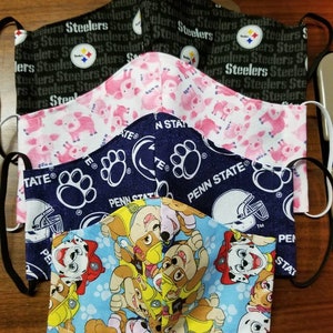 May include: A stack of four fabric face masks. The top mask is black with the repeated text "Steelers". The second mask is pink with a white pig pattern. The third mask is blue with a white paw print and the text "Penn State". The bottom mask is blue with a white paw print and a cartoon pattern of puppies.