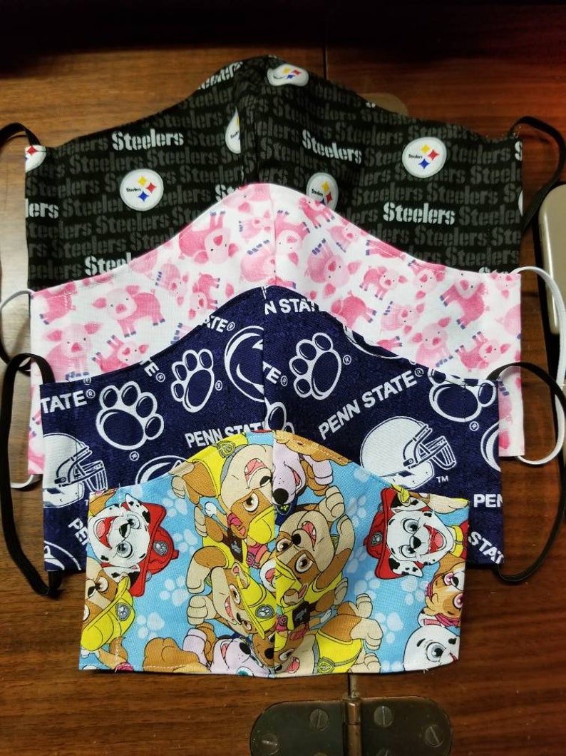 May include: A stack of four fabric face masks. The top mask is black with the repeated text "Steelers". The second mask is pink with a white pig pattern. The third mask is blue with a white paw print and the text "Penn State". The bottom mask is blue with a white paw print and a cartoon pattern of puppies.
