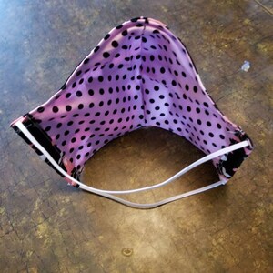 May include: A pink and black polka dot face mask with white elastic ear loops. The mask is folded in half and the inside of the mask is visible.