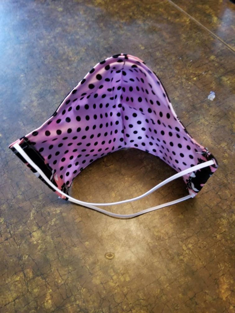 May include: A pink and black polka dot face mask with white elastic ear loops. The mask is folded in half and the inside of the mask is visible.