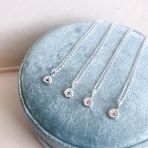 Może przedstawiać: Cztery delikatne srebrne naszyjniki z małymi, okrągłymi wisiorkami. Każdy wisiorek ma inny kolor kamienia szlachetnego: niebieski, zielony, różowy i pomarańczowy. Naszyjniki leżą na jasnoniebieskiej aksamitnej powierzchni.