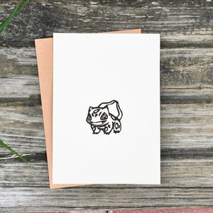 Könnte beinhalten: Eine weiße Grußkarte mit einer schwarzen Bulbasaur-Illustration, die auf einem braunen Umschlag liegt. Karte und Umschlag befinden sich auf einer verwitterten Holzoberfläche, mit grünem Gras im Hintergrund.
