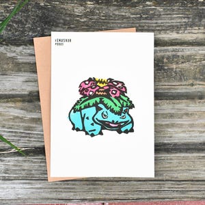 Könnte beinhalten: Eine weiße Karte mit einer blau, grün und rosa Illustration von Venusaur. Die Karte liegt auf einem braunen Umschlag, mit dem Text "VENUSAUR #0003" oben links. Der Hintergrund ist eine verwitterte Holzoberfläche.