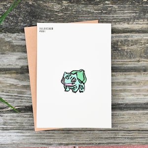 Könnte beinhalten: Eine weiße Karte mit einer Bulbasaur-Illustration und dem Text "Bulbasaur #001". Sie wird von einem braunen Umschlag begleitet, der auf einer rustikalen Holzoberfläche liegt.