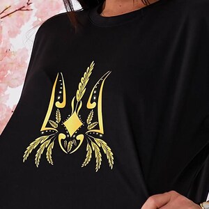 Puede incluir: Camiseta negra con un s&iacute;mbolo de tridente ucraniano bordado en oro.