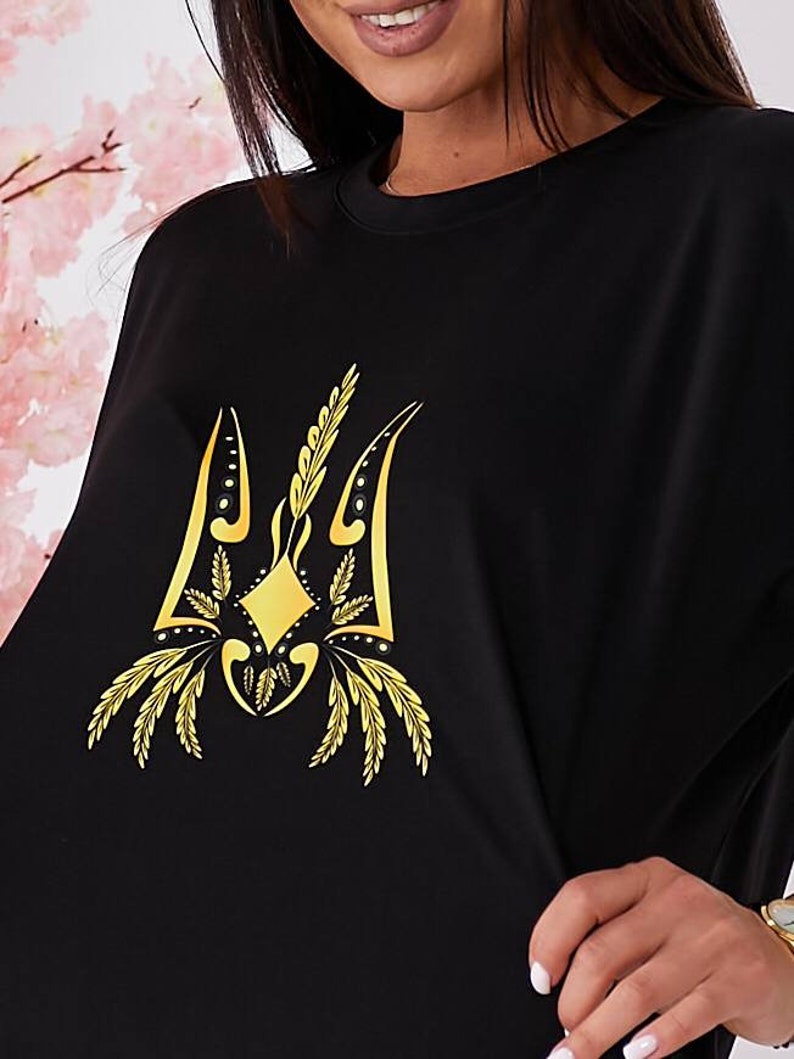 Puede incluir: Camiseta negra con un s&iacute;mbolo de tridente ucraniano bordado en oro.