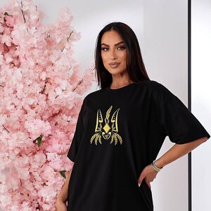 Puede incluir: Camiseta negra con un s&iacute;mbolo de tridente ucraniano bordado en oro.
