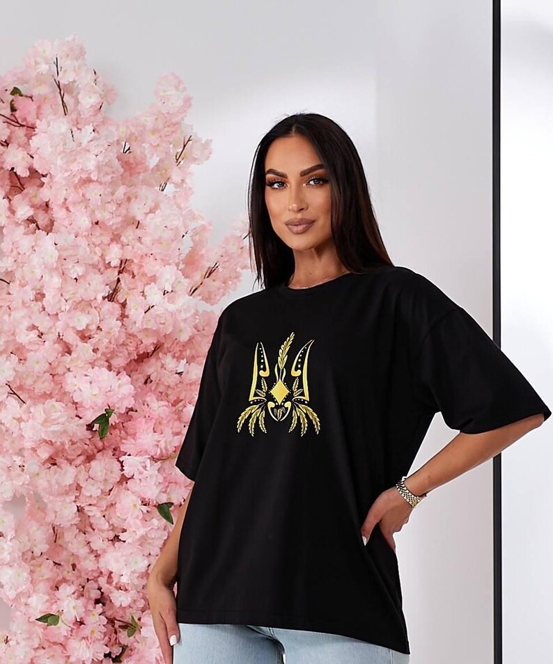 Puede incluir: Camiseta negra con un s&iacute;mbolo de tridente ucraniano bordado en oro.